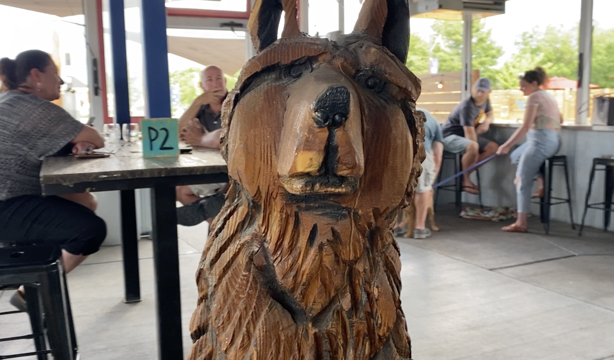 This is a wooden statue of a dog at Romer's K9 Club Taproom. Behind the statue are people drinking beer.