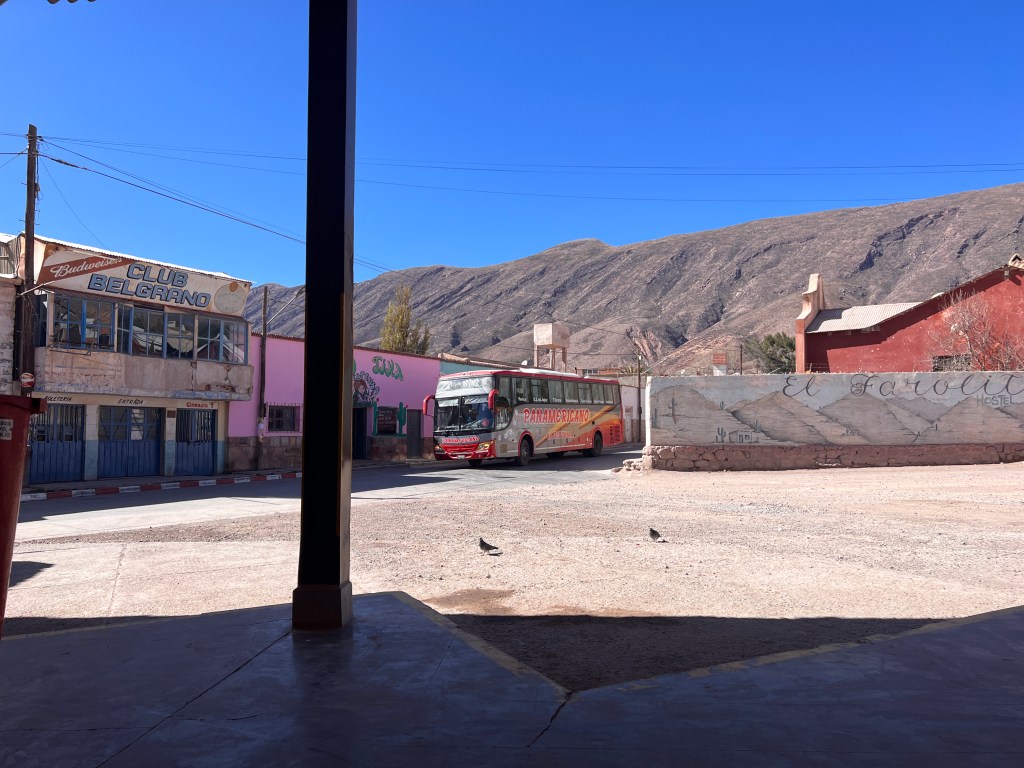A bus is pulling into the Tilcara bus station in the north of Argentina. There is a large gravel lot that is empty. Concrete platforms can be seen in the foreground.