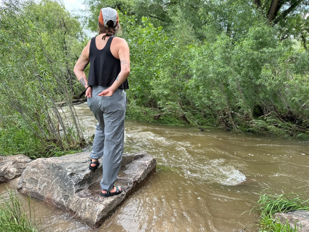 Hiking Gear List for Beginners: Nicki stands on a rock with her back to the camera, wearing a black tank, a cap, and grey Kuhl hiking pants.