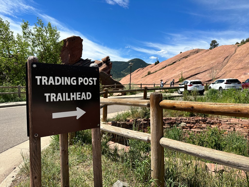 There is a wooden sign that reads "Trading Post Trail Ahead" and the arrow points to the right, beyond the parking lot.