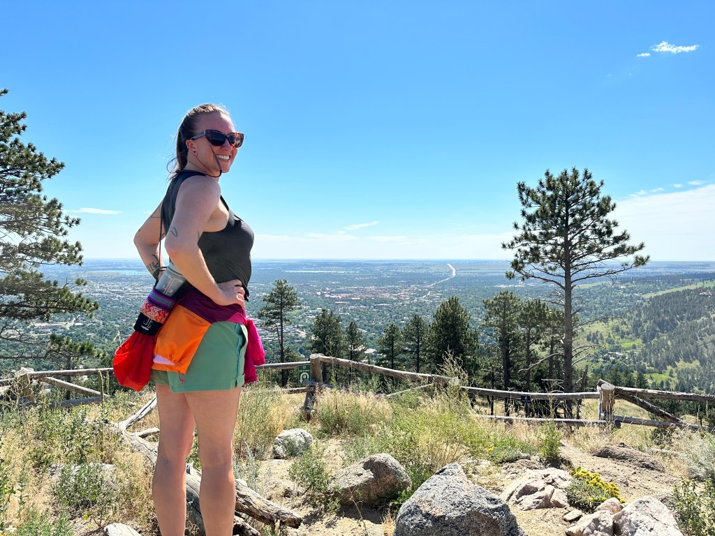 Nicki stands with her back to the camera, but turned around to face the camera. She is on the left side of the photo. In the background there are trees and a wooden fence. Down below is the city of Boulder. She is on the peak of the Anemone Trail at Anemone Point.