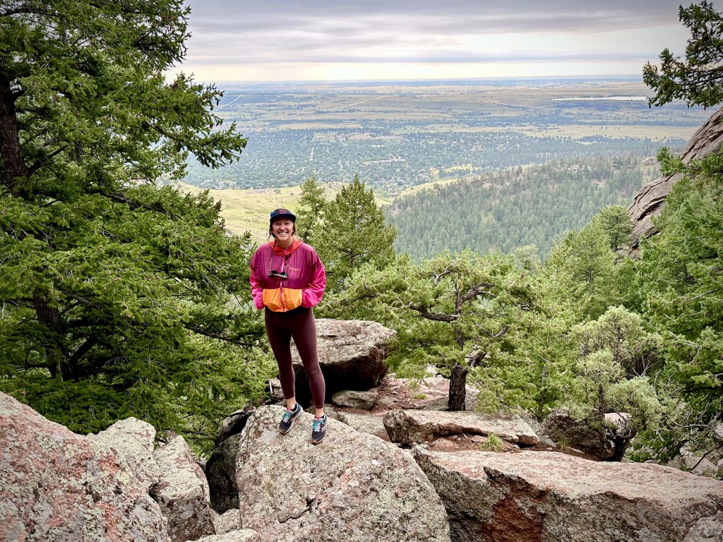 Nicki stands in the center of the photo, smiling. She is standing on a large rock with trees around her. In the background is more trees, parts of the city of Boulder, and the horizon in the far distance.