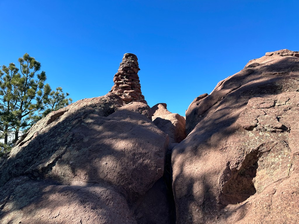 There are several very large boulders. On top of the boulders is a stack of smaller rocks. on top of that is a plaque. This marks the official peak of Green Mountain.