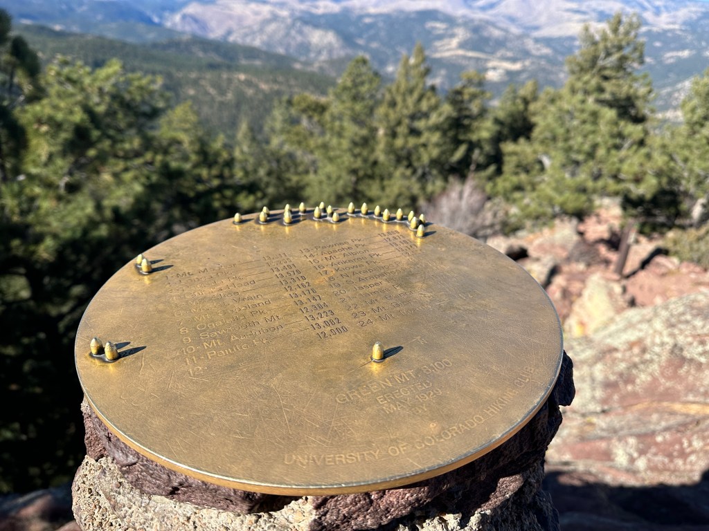This is a circular plaque that sits at the very summit of Green Mountain. There are several mountain peak names carved into this plaque with their elevations listed. You can see the mountainous terrain in the distance behind the plaque.