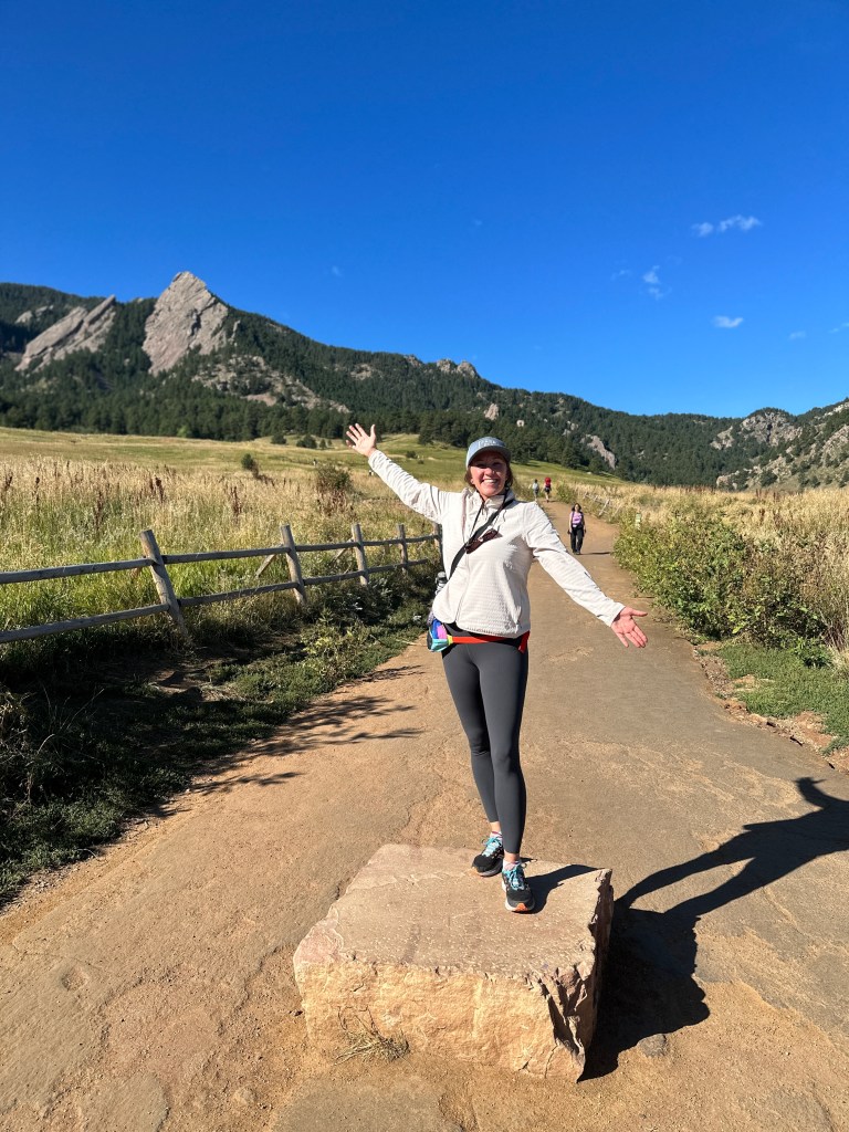Nicki stands at the Chautauqua Trailhead. She is standing on top of a rock. Behind her in the distance are the flatirons. She has her hands raised.