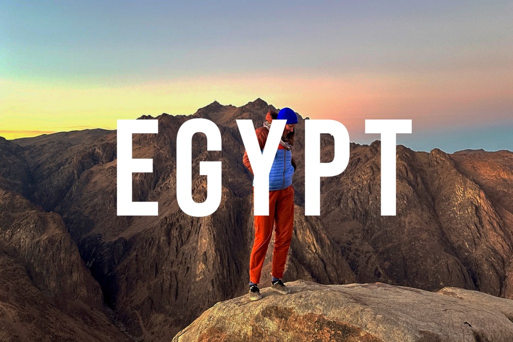 This is an image of Nicki standing on Mt Sinai at sunrise. She is wearing a Cotopaxi jacket and pants and a blue beanie. Behind her is a mountain and a blue and orange sunrise. Over the top of the image is the word: EGYPT