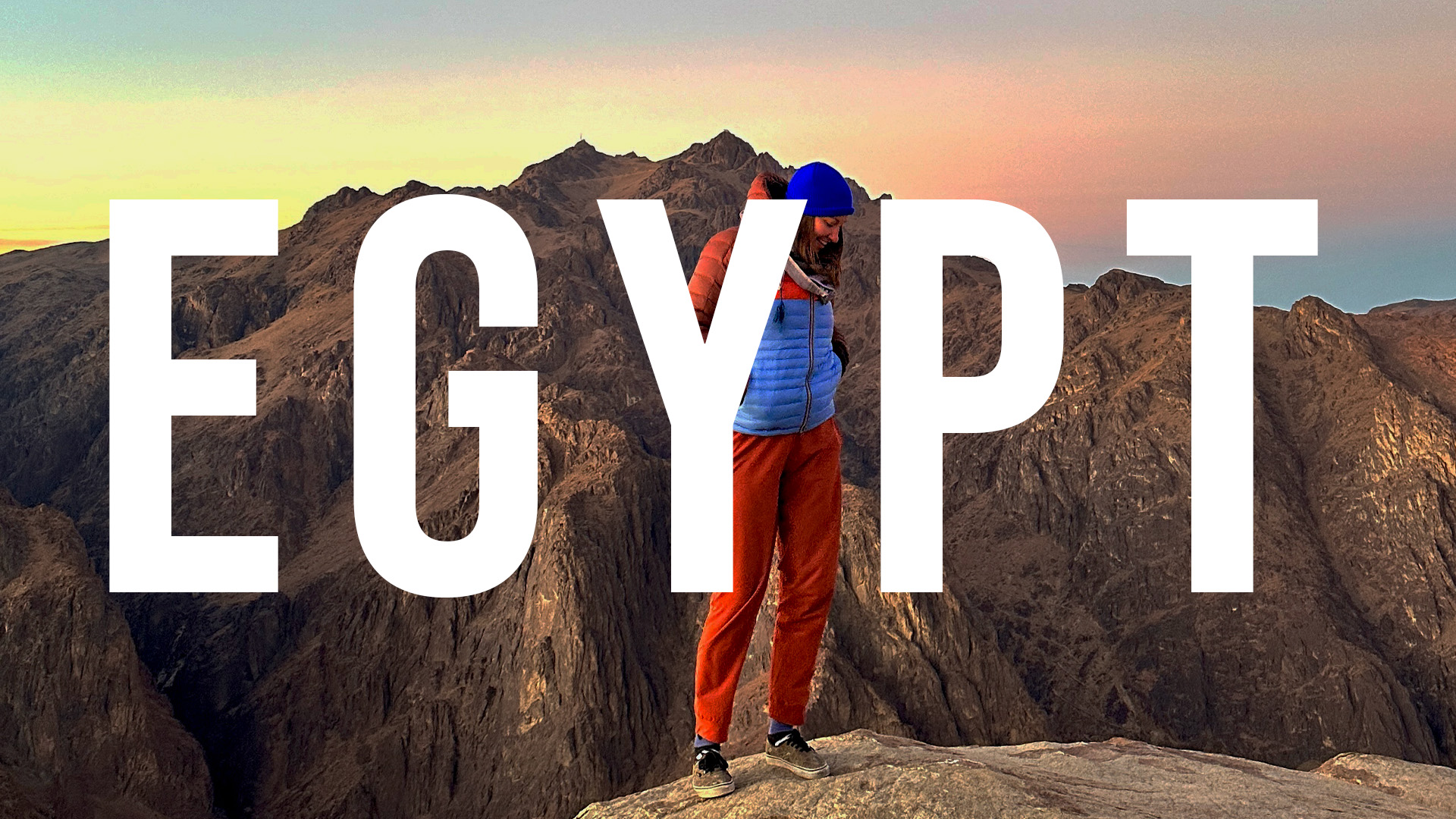This is an image of Nicki standing on Mt Sinai at sunrise. She is wearing a Cotopaxi jacket and pants and a blue beanie. Behind her is a mountain and a blue and orange sunrise. Over the top of the image is the word: EGYPT