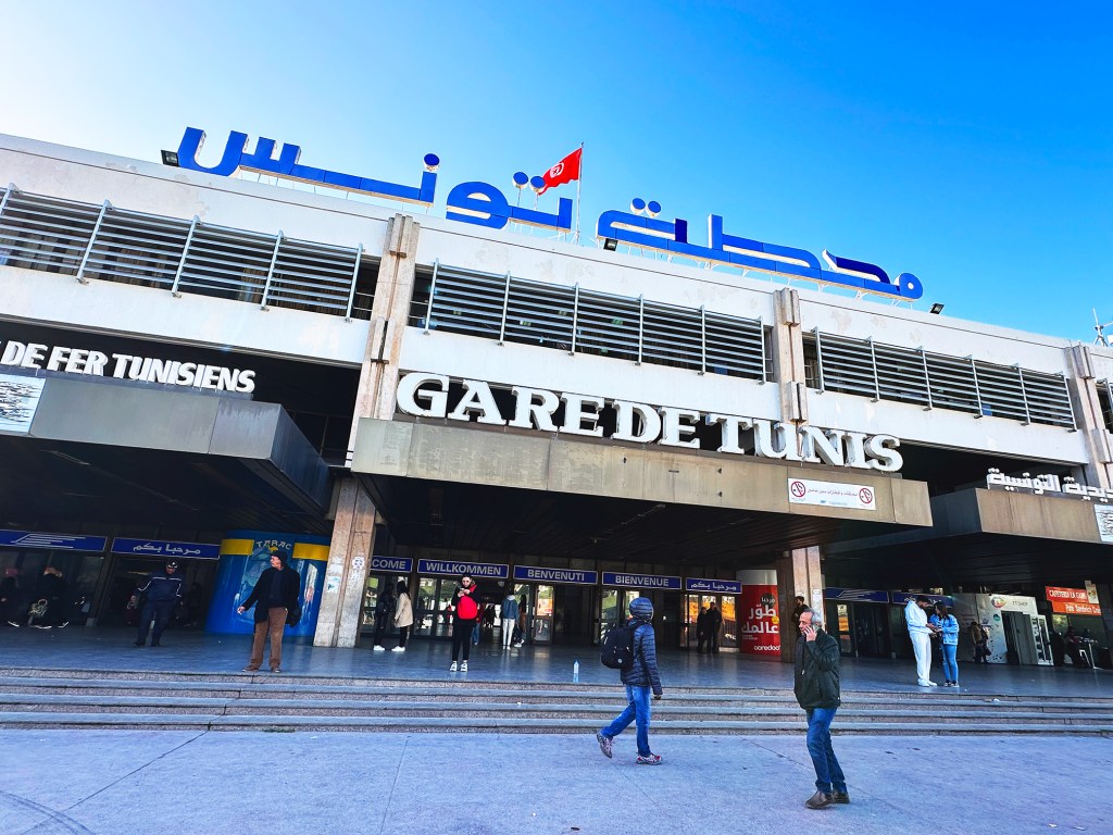 This is the front of the Tunis train station. It has the words in Arabic on the top in blue. In white it says "Gare de Tunis." There is a Tunisian flag on the top.