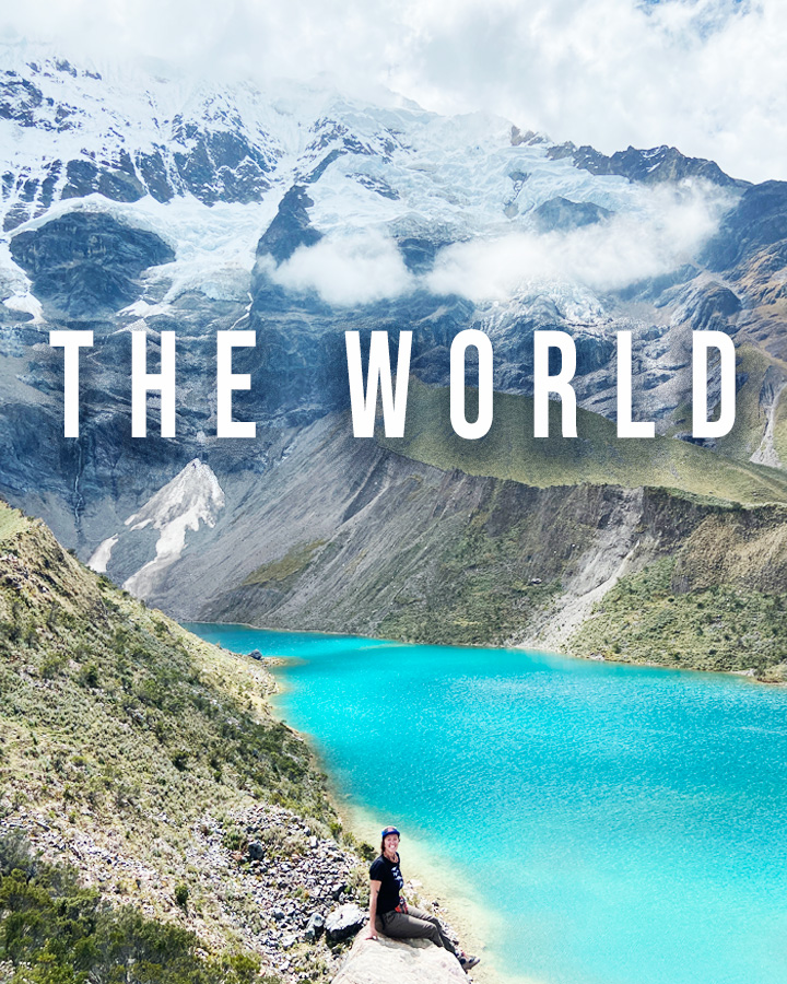 Nicki sits on a rock at the bottom of the photo. Behind her is the stunningly blue Humantay Lake in Peru, with the Salkantay mountain behind her in the distance. Superimposed over the photo is the word "THE WORLD"
